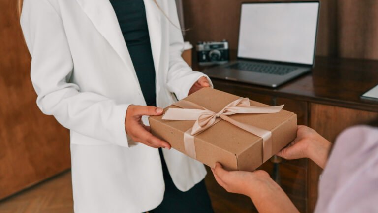 hands exchanging gift box wrapped in craft paper with beige ribbon. woman in white blazer receives present indoors near laptop and retro camera on wooden cabinet